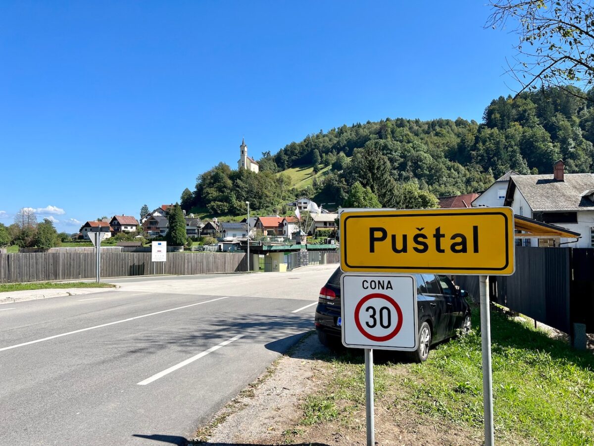 A sign announcing the entrance to the small town of Postal in Slovenia with the Church of the Holy Cross in the background