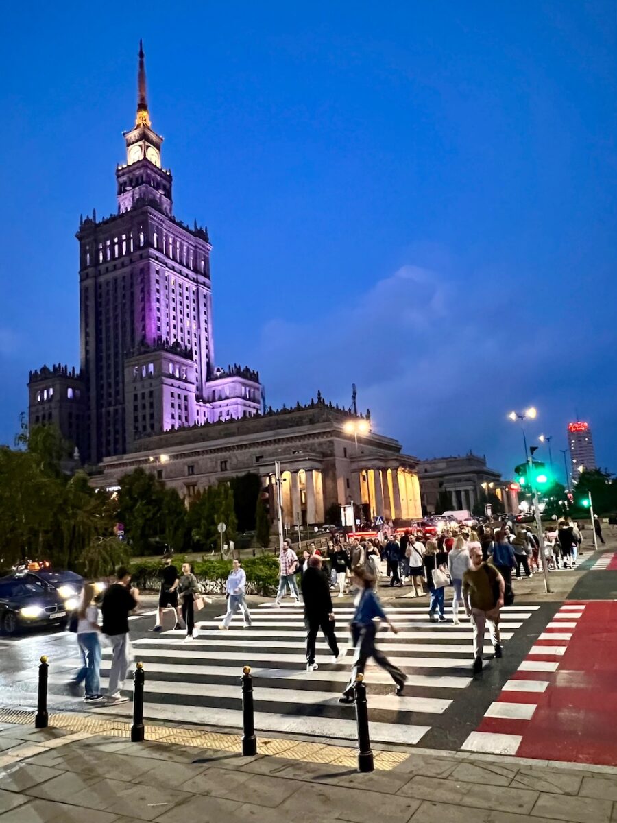 The Palace of Culture and Science building in Warsaw Poland lit up in purple lights at night with pedestrians crossing a crosswalk